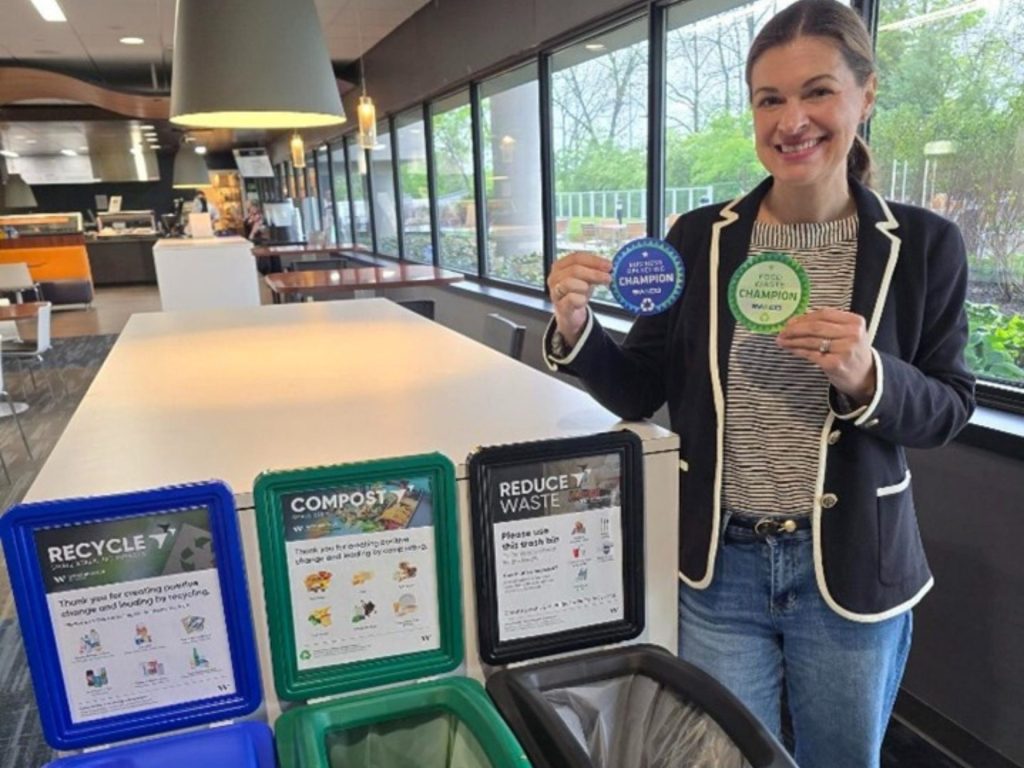 A woman holding both the SWACO recycling and compost champion badges standing next to recycling, compost, and landfill bins.