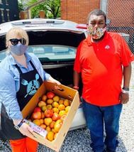 Two people standing in front of a car. One is holding a box of apples.