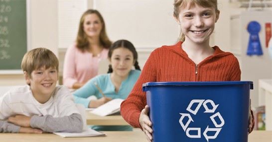 A girl at the front of her class holding a recycling bin.
